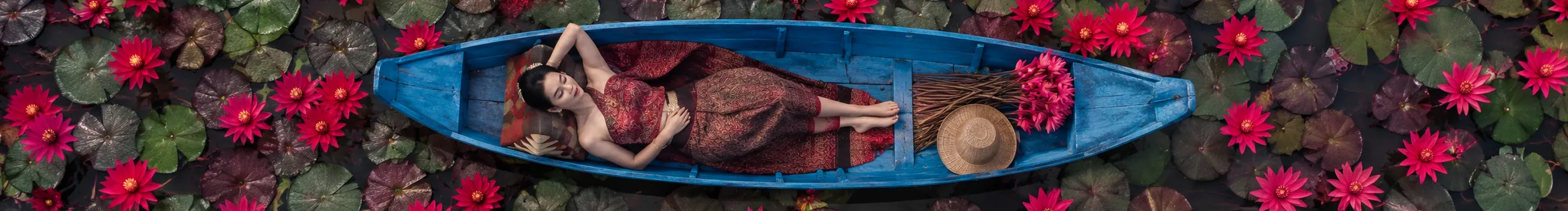 Woman in traditional Thai dress lying in a boat surrounded by red lotus flowers, Nakara Villas and Glamping