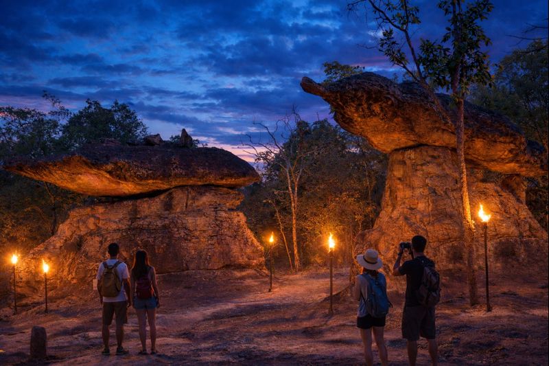 Tourists at Phu Phra Bat Historical Park in Udon Thani viewing ancient mushroom-shaped rock formations at blue hour with torchlight